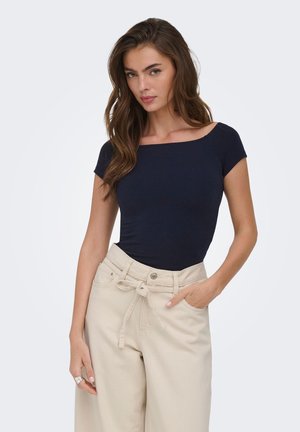 Kobieta o długich brązowych włosach, ubrana w dopasowany granatowy krótki top z krótkim rękawem i beżowe spodnie z wysokim stanem oraz paskiem do wiązania, stoi z jedną ręką w kieszeni.