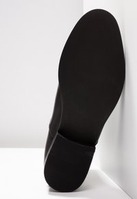 Semelle de chaussure en cuir noir avec talon texturé et avant-pied lisse, reposant verticalement contre un coin mur-sol blanc.