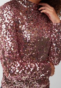 Top con paillettes in rosa oro, con collo alto, maniche lunghe e superficie testurizzata. Presenta paillettes circolari sovrapposte in diverse tonalità.