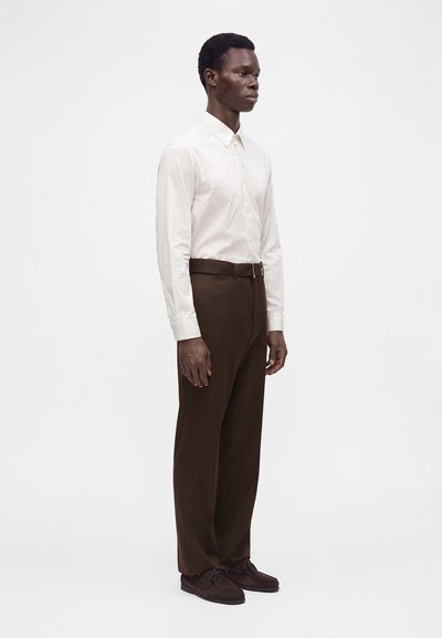 Homme debout de profil portant une chemise blanche à manches longues, un pantalon marron avec une ceinture et des chaussures marron foncé à lacets sur un fond blanc uni.