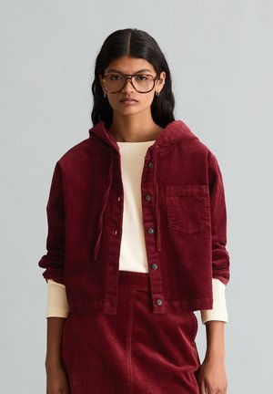 Femme aux longs cheveux noirs et lunettes, portant une veste en velours côtelé bordeaux avec des boutons et une jupe assortie sur une chemise crème.