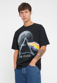 Musta puuvillapaita, jossa on pyramidi sateenkaaren väriin liittyvä graafinen kuva. Lyhyet hihat, rento istuvuus ja teksti "PINK FLOYD" valkoisella.