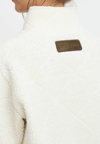 Giacca bianca in pile con un motivo trapuntato testurizzato. Presenta un'etichetta di tessuto marrone sul retro vicino al collo.