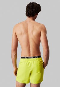 Neon gele zwembroek met een elastische zwarte tailleband waarop het "Calvin Klein" logo staat. De shorts zijn knie-lengte en hebben een achterzak.