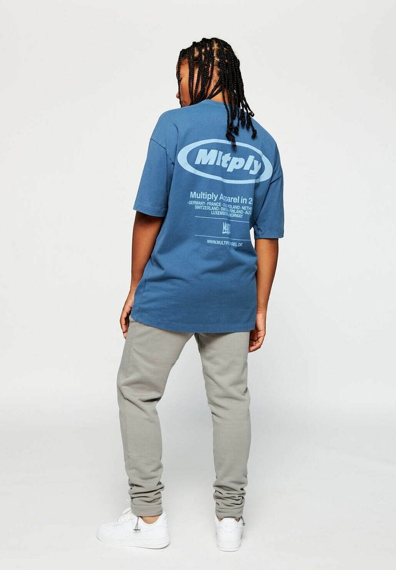 T-shirt en coton bleu avec un logo dans le dos, associé à un pantalon de jogging gris clair. Des baskets blanches complètent la tenue.