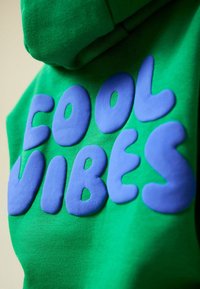 Zöld kapucnis pulóver, nagy, kiemelkedő lila betűkkel, amelyek a "COOL VIBES" feliratot alkotják. Simap felületű, kenguruzsebbel és kapucnival.