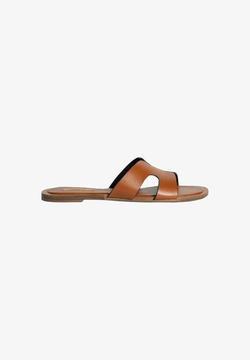 Sandal de piel marrón con una cinta ancha y curva y un detalle cortado. Suela plana con una base color tostado y un diseño minimalista.