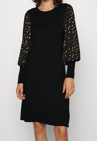 Svart klänning med långa, puffiga ärmar som har guldprickar. Tillverkad av mjukt tyg med rund halsringning och en avslappnad passform.