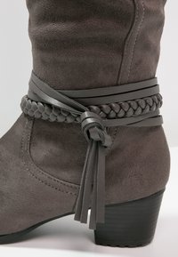 Botte en suède gris foncé à talon moyen avec des lanières en cuir tressé et lisse enroulées autour de la cheville, nouées en un nœud avec des pampilles.