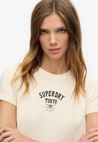 Kratka majica s kratkimi rokavi kremne barve z black "SUPERDRY TOKYO" grafiko, ki vključuje diamante in žarke. Mehka tkanina z diskretno teksturo.