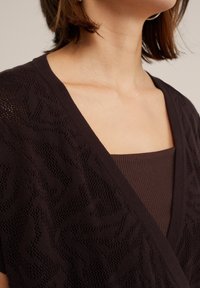 Tmavě hnědý pletený cardigan s texturovaným vzorem, s hlubokým výstřihem do V, nošený přes odpovídající žebrovaný top.