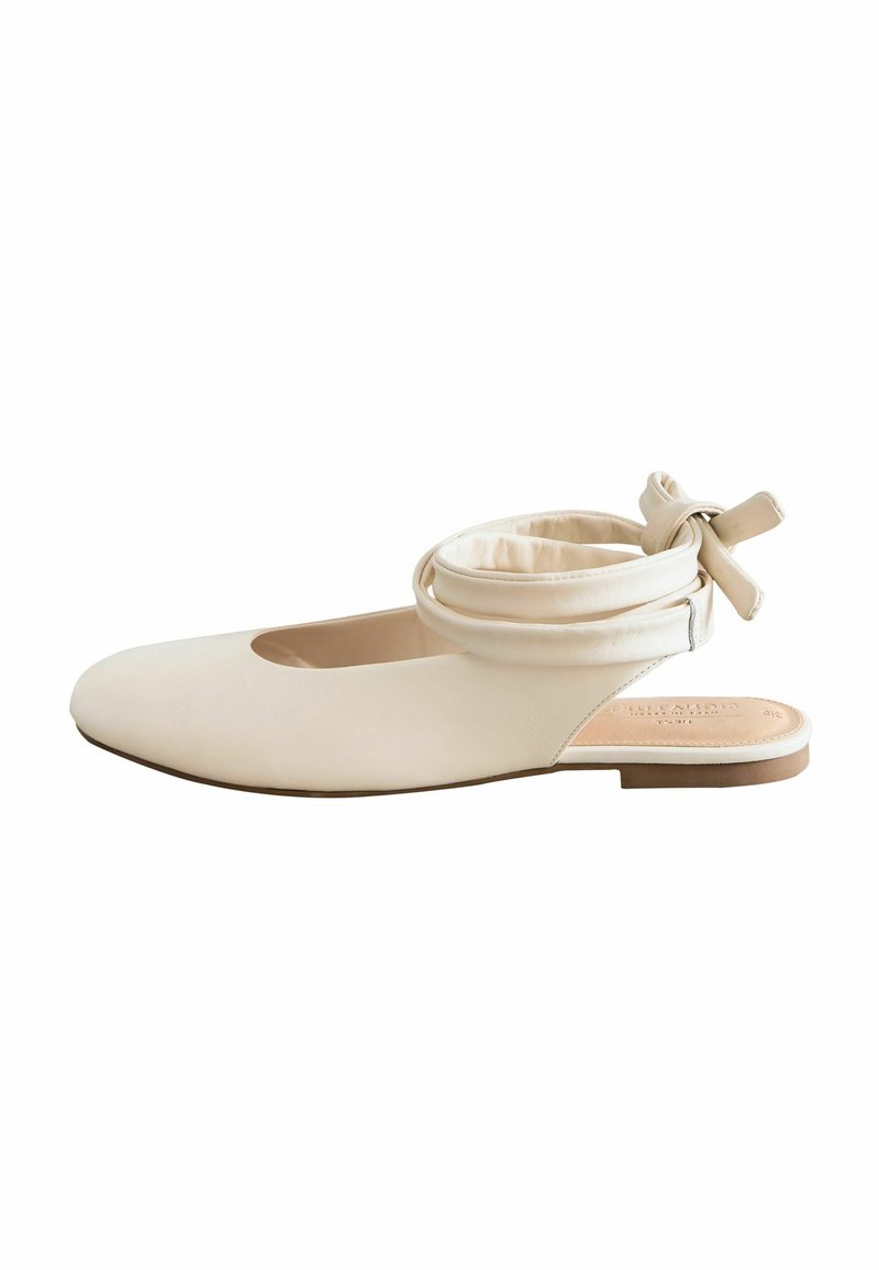 Flat beige shoe with a rounded toe, featuring a cut-out detail and a soft, wrapped ankle strap with a bow. Smooth leather finish and minimal design.