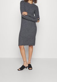 Robe tricotée grise à manches longues, texture côtelée, et silhouette ajustée. Comprend un détail de boutons sur l'épaule et des fentes sur les côtés.