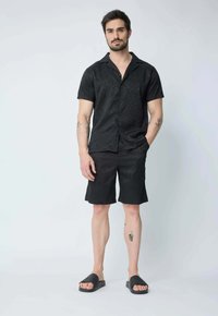 Chemise noire à manches courtes et boutons, avec un motif texturé en vagues et un short assorti, complétée par des sandales glissantes noires.