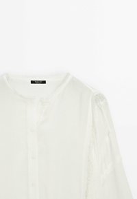 Blouse blanche à manches longues avec ouverture boutonnée à l'avant et détails en dentelle sur les épaules et les manches.