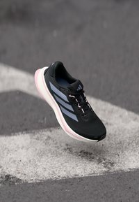 Zwarte sportieve schoen met een textuurmesh bovenwerk, roze en witte zool, en drie grijze strepen aan de zijkant. Lichtgewicht ontwerp, geschikt voor hardlopen.