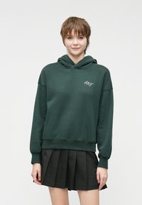 Dark green hoodie made of soft cotton blend, featuring a front pocket and ribbed cuffs. Paired with a black pleated skirt.