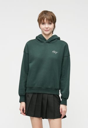 Hoodie vert foncé en mélange de coton doux, avec une poche avant et des poignets côtelés. Associé à une jupe plissée noire.