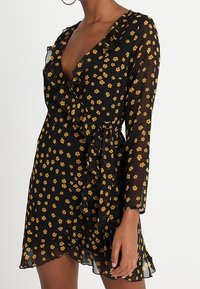 Černé šaty s dlouhými rukávy, výstřihem do V a volánkovým detailem na přední části. Obsahují žlutý květinový potisk a stáhnutý pas, s volným lemem.