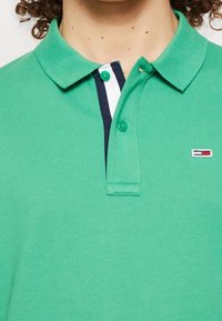 Zelená polo tričko vyrobené z texturované látky. Obsahuje tříknoflíkovou légu, kontrastní námořnickou a bílou pruh uvnitř límce a malé logo.