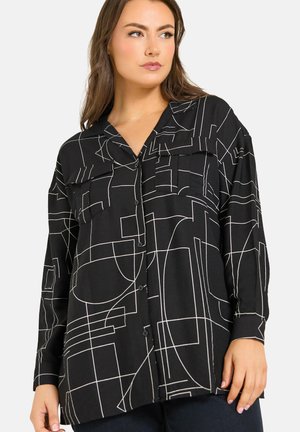Kvinde iført en sort skjorte med knapper, med hvide abstrakte geometriske linjemønstre, lange ærmer og to brystlommer.