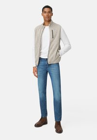Lichtgrijze fleece vest met een hoge kraag, rits aan de voorkant en zijzakken; gedragen over een wit shirt met lange mouwen en blauwe jeans met bruine laarzen.