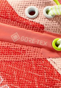 Rød og rosa syntetisk skomateriale med et teksturert mønster, hvite øyer, og et synlig GORE-TEX-merke i et fremtredende område.