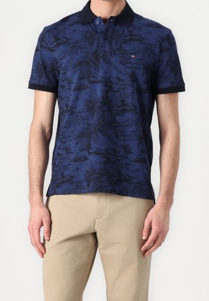 Hombre con camisa polo azul marino con estampado tropical negro y pantalones beige, de pie frente a un fondo blanco liso.