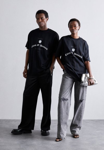 Sorte bomuld t-shirts med trykt tekst, parret med løse sorte bukser og blegede grå jeans med huller, akkompagneret af en sølvfarvet clutch. 