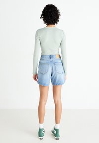 Svetlo zelena majica s dugimi rukavi, denim kratke hlače ohlapnega kroja, ki imajo dve zadnji žepi. Model nosi zelene visoke superge z belimi detajli.