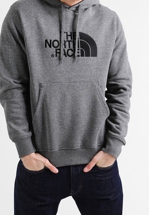 Mężczyzna ubrany w szarą bluzę z kapturem The North Face z przednią kieszenią oraz ciemnoniebieskie dżinsy, ręce włożone do kieszeni dżinsów.