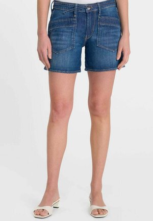 Donna che indossa pantaloncini di jeans blu con tasche frontali e sandali bianchi con tacco basso e punta aperta, in piedi davanti a uno sfondo semplice.