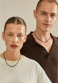 Czarne naszyjniki choker na dwóch modelkach; jedna w jasnym swetrze, a druga w ciemnej koszulce polo. Neutralne tło z miękkim oświetleniem.