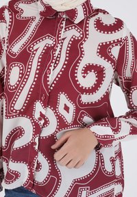 Bordeaux- und weiße gemusterte Bluse mit Knopfleiste, Kragen und filigranen, wirbelnden Designs mit kleinen Punkten. Glatte Stofftextur.