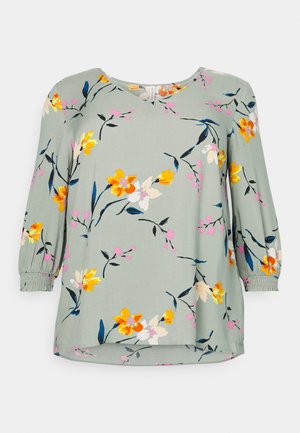 Blouse florale gris clair avec des motifs de fleurs jaunes, roses et bleues. Dotée de manches trois-quarts et de poignets froncés. Tissu lisse.