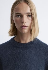 Navyblauer, gestrickter Pullover mit rundem Ausschnitt, der eine weiche, flauschige Textur aufweist. Das Model hat kurze Haare und trägt goldene Creolen.