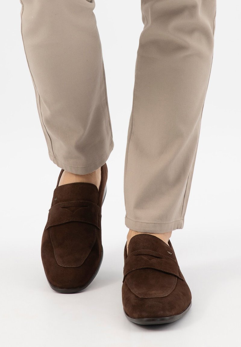 Personne portant un pantalon beige et des mocassins en daim marron foncé debout sur un fond clair.