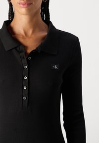 Melna ribota garām piedurknēm polo krekls ar apkakli, kam ir pogu aizdare un neliels CK logotips uz krūtīm.