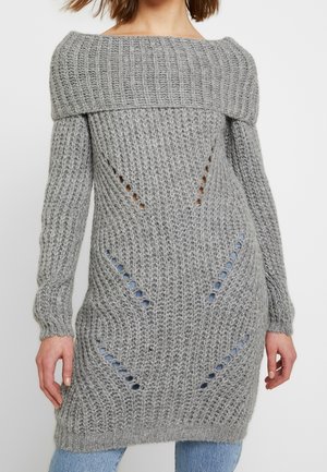 Woman wearing a long gray knitted off-shoulder sweater with textured patterns and small open holes, paired with blue jeans.