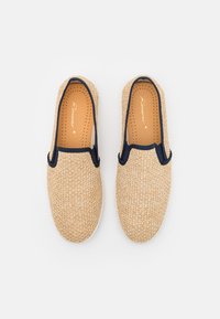 Beige geweven instappers met een gestructureerd oppervlak, marineblauwe randen en geperforeerde binnenzool, die een eenvoudig, functioneel ontwerp laten zien.