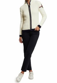 Kabelstickad krämfärgad cardigan med dragkedja framtill, marinblå detaljer och logotypmärke. Såld tillsammans med mörka byxor och vita sneakers.