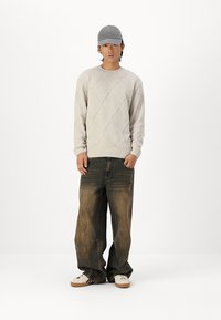 Light gray knitted sweater with diamond pattern, paired with loose, faded dark denim jeans and white sneakers. The model wears a gray cap.