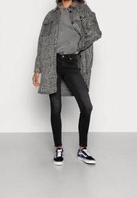 Abrigo largo a cuadros en blanco y negro, camiseta gris, jeans skinny negros y zapatillas negras con toques en azul y púrpura. Conjunto casual en capas.