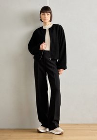 Veste noire texturée zippée, haut crème côtelé, pantalon noir taille haute et sneakers beiges. Surfaces lisses et couleurs contrastées.