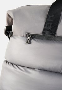 Gray puffy backpack featuring a smooth texture, zipper pocket, and a metallic "B" zipper pull. Includes a thick black strap with branding.