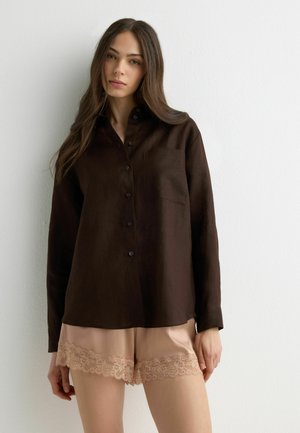 Junge Frau mit langen braunen Haaren, die ein dunkelbraunes Hemd mit Knöpfen und beigen Shorts mit Spitzenbesatz vor einem schlichten weißen Hintergrund trägt.