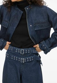 Tumši zils džinsa žakete ar kroplētu dizainu, kas piedāvā divas priekšējās kabatas un sudraba rāvējslēdzēju. Kombinēts ar augsta vidukļa biksēm un melnu topu.