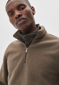 Brun fleece pullover med kvart lynlås har en tekstureret overflade, høj krave og en sølv lynlås. Et mørkt mønstret tørklæde bæres under.