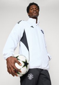 White athletic jacket with black accents, zippered front, and logo. Grey pants with club emblem. Model holds a white and black soccer ball.