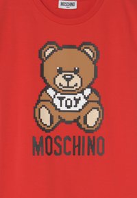 T-shirt rouge avec un graphique en pixels d'un ours en peluche brun, portant le mot "TOY" sur sa chemise et le texte "MOSCHINO" en gras noir en dessous. Matière en coton doux.